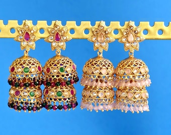 May include: Two pairs of gold-tone jhumka earrings. The left pair features pink and green stones with matching beads. The right pair features light pink stones and beads. Each earring has a floral stud and a double-tiered bell shape.