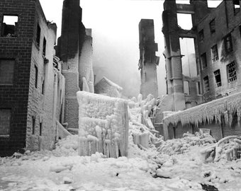 May include: A black and white photograph of a building covered in ice. The building appears to be damaged and has a brick exterior. The ice is hanging from the building in long icicles.