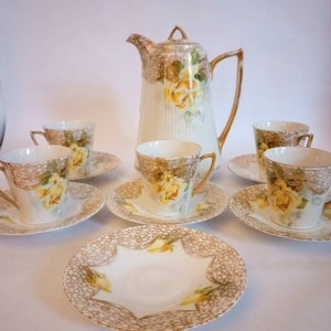 May include: A vintage tea set with a white background and gold accents. The set includes a teapot, four cups, and four saucers. The cups and saucers are decorated with yellow roses and a delicate lace pattern.