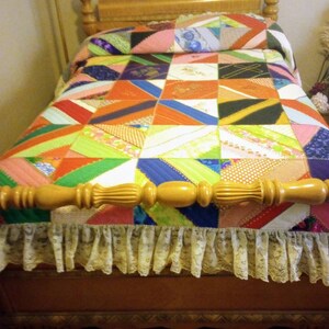 May include: A colorful patchwork quilt with a variety of fabrics and patterns, including stripes, polka dots, and floral designs. The quilt is laid out on a wooden bed frame with a white lace trim.