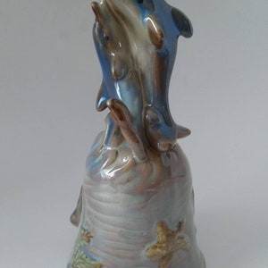 May include: A ceramic bell with a blue and brown glazed finish, featuring two dolphins jumping out of the water. The bell is decorated with a starfish and other sea life.