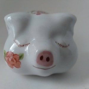 May include: White ceramic piggy bank with closed eyes and a pink snout. The piggy bank has a floral design with pink roses.