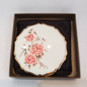 May include: A white porcelain compact mirror with a gold trim and a floral design featuring pink roses. The mirror is in a brown box with a black velvet lining.