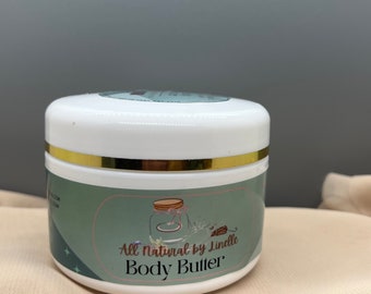 Op de afbeelding: Een witte pot met een gouden deksel en een groen label met de tekst "All Natural by Lindle Body Butter".