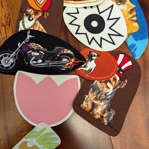 May include: A collection of iron-on patches featuring various designs, including a hot dog, a dog wearing a hat, a motorcycle, a flower, a dog wearing a hat, and a cartoon character.