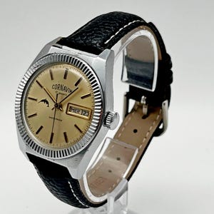 May include: A vintage Cornavin wristwatch with a gold-toned dial, silver-colored fluted bezel, and a black textured leather strap with white stitching. The dial features a dolphin logo, the text CORNAVIN 17 jewels SHOCK PROOF, and a day-date window.