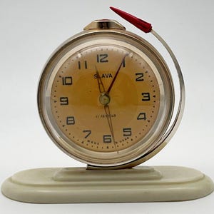 May include: A vintage Slava desk clock with a round gold-tone metal frame and a light orange face featuring black Arabic numerals. The clock includes a red pointer arm that curves around the side and sits on an off-white oval base. Text reads Slava 11 Jewels Made in USSR.
