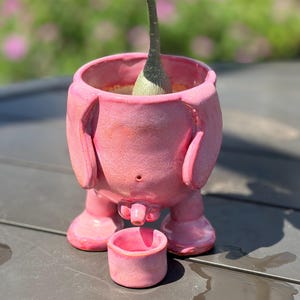 May include: A pink ceramic planter shaped like a stylized figure with two legs and two side appendages. The planter features a small central protrusion and includes a separate, smaller matching pink cylindrical cup placed in front of the base.