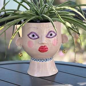 May include: A whimsical, hand-painted ceramic planter shaped like a face, with a light pink base color. The face features purple eyes, pink cheeks, and red lips. The planter is adorned with a blue necklace and yellow and black earrings. The planter holds a green and white plant.