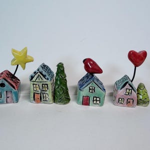 May include: A set of four small ceramic houses with hand-painted details. One house features a yellow star on a wire, another has a red bird on the roof, and a third has a red heart on a wire. Two small green ceramic trees accompany the houses.