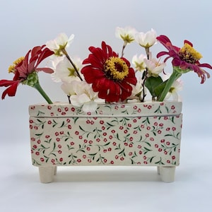 May include: A rectangular ceramic vase with a floral pattern, holding a bouquet of red and white flowers. The vase is cream-colored with a repeating pattern of small red flowers and green leaves. The vase is 6 inches long and 3 inches wide.