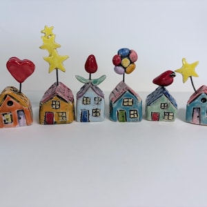 May include: A set of six decorative ceramic houses, each with a different topper. The houses are painted in various colours and feature windows and doors. Toppers include a red heart, yellow stars, a red tulip, colourful spheres, a red bird, and a yellow star.