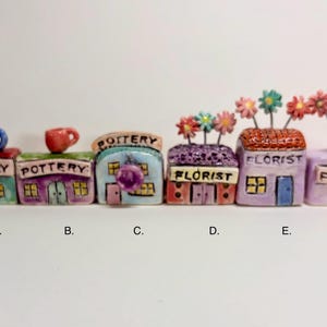 May include: A set of six miniature ceramic buildings labeled A through F. The structures include pottery shops with small mugs on top and florist shops featuring wire-stemmed flowers. Each building is hand-painted in pastel colors with text.