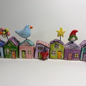 May include: A row of seven small, colorful ceramic houses. The houses feature various roof colors, including blue, purple, pink, and green. Decorative elements include a bird, a flower, a yellow star, red hearts, and small gnome figures.