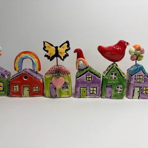 May include: A set of six small, colorful ceramic house figurines arranged in a row. Each house features a unique topper, including a flower, a rainbow, a butterfly with a heart, a gnome, a red bird, and another flower, all on wire stems.