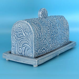 May include: A light blue ceramic butter dish with a lid and a matching rectangular base. The dish features a detailed embossed floral and snowflake design. The lid has a small, rounded handle.
