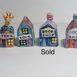 May include: Four small ceramic miniature buildings. The first is an orange and blue toy shop with a giraffe on top. The second is a blue toy shop. The third is a blue book shop with a pink roof. The fourth is a purple book shop with a pink roof.