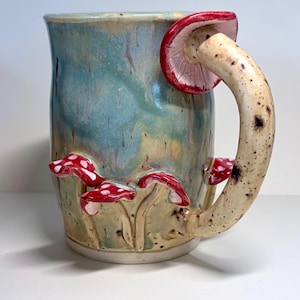 May include: A ceramic mug with a blue and green glazed body featuring three-dimensional red and white spotted mushrooms at the base. The handle is shaped like a long mushroom stalk with a red cap attached to the top rim of the mug.