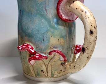 May include: A ceramic mug with a blue and green glazed body featuring three-dimensional red and white spotted mushrooms at the base. The handle is shaped like a long mushroom stalk with a red cap attached to the top rim of the mug.
