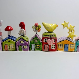 May include: A set of seven small, colorful ceramic house figurines arranged in a row. Each house features unique hand-painted details including a white rabbit, a red mushroom, a gnome, a flower, a yellow bird, yellow stars, and a red heart.