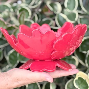 May include: A bright red ceramic flower-shaped bowl, held in a hand. The bowl has multiple petals and a glossy finish. The background features green and white foliage, creating a natural contrast.
