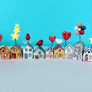 May include: A collection of fourteen miniature ceramic houses arranged in a row. Each house features a unique color and a wire topper displaying various shapes including red birds, yellow stars, red hearts, and colorful clusters of spheres.