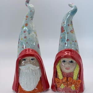May include: Two ceramic gnome figurines. One has a white beard and red hood, the other has blonde braids and a red hood. Both have tall, speckled, light blue hats with a curved top. The gnomes are painted with bright colors.