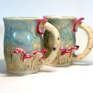 May include: Two ceramic mugs featuring a light blue and cream glazed finish with speckled textures. Each mug has a curved handle and is decorated with small, raised red mushrooms with white spots attached to the exterior surface of the body.