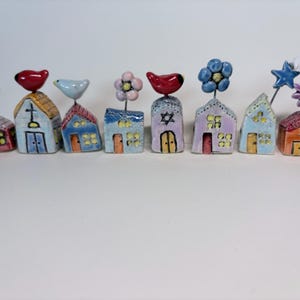 May include: A collection of eight small, hand-painted ceramic house figurines arranged in a row. Each house features unique details like birds, flowers, stars, or religious symbols on top, with colorful painted windows and doors on the facades.