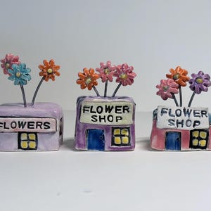 May include: Three small ceramic structures shaped like shops, each featuring a sign reading "FLOWERS" or "FLOWER SHOP" and topped with wire stems holding colorful clay flowers in shades of pink, blue, orange, and purple with yellow centers.