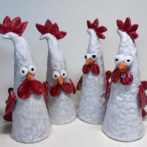 May include: Four ceramic chicken figurines with white textured bodies, red combs and wattles, orange beaks, and large round eyes. Each figure has a tall, tapered shape with a wide base, standing upright with distinct red tail feathers visible.