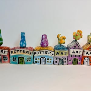 May include: A set of six miniature ceramic buildings arranged in a row. Three buildings are labeled POTTERY and three are labeled ART. Each structure features a unique colorful roof, a small door, windows, and a distinct decorative topper.