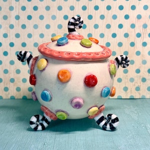 May include: A round ceramic lidded jar with a cream-colored base, decorated with raised, multi-colored polka dots in shades of red, orange, yellow, blue, and green. It features three black and white striped curled feet and a matching lid handle.