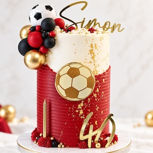 Puede incluir: Un pastel de varios niveles con base roja estriada y parte superior blanca, decorado con un emblema de balón de fútbol dorado, un adorno con el nombre Simon, el número 40 dorado, una vela dorada y esferas negras, rojas y doradas con un balón.