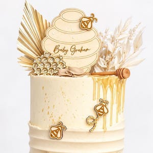 May include: A two-tiered cream-coloured cake decorated with gold honey drip, honeycomb accents, and bee-shaped toppers. A wooden cake topper reads Baby Graham. The cake features dried palm leaves, white floral sprigs, and a Winnie the Pooh cookie.