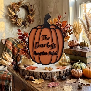 May include: A decorative orange pumpkin-shaped sign with a black outline and the black text The Darby's Pumpkin Patch. The sign is displayed on a circular wood slice base surrounded by small decorative pumpkins and dried autumn foliage.