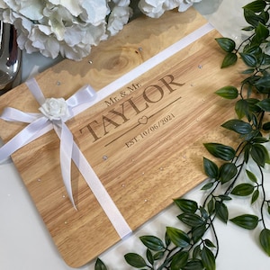 以下が含まれることがあります： 「Mr. & Mr. TAYLOR EST. 10/06/2021」と刻まれた木製のまな板。小さなラインストーンが付いています。白いリボンと白いバラがボードに付いています。白い花と緑の葉がボードを囲んでいます。