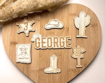 Puede incluir: Decoraciones de madera con temática de vaquero sobre una tabla de madera en forma de corazón. Incluye una placa de sheriff con el número 3, un sombrero de vaquero, un cactus, el nombre "GEORGE", un letrero de "SE BUSCA", una bota de vaquero y un letrero de "OESTE SALVAJE".