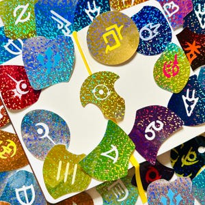 Op de afbeelding: Een verzameling kleurrijke, glinsterende stickers in verschillende vormen, waaronder cirkels en schilden. De stickers tonen symbolen in wit, geel en roze tegen achtergronden van blauw, groen, goud en rood. Een gele stok is ook zichtbaar.