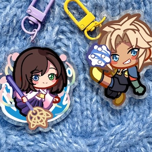 May include: Two anime-style acrylic keychains with metal clasps. One keychain features a character with brown hair and a purple outfit, holding a purple staff. The other keychain depicts a character with blonde hair and blue eyes. Both keychains are on a blue knitted background.