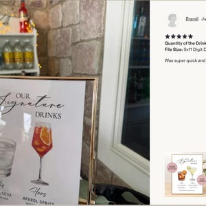 Pu&ograve; includere: Un cartello rettangolare con cornice dorata che riporta il testo Our Signature Drinks, con illustrazioni di un tequila soda e un Aperol spritz. Il cartello elenca His Tequila Soda con lime e Hers Aperol Spritz con arancia, in una cornice di vetro.
