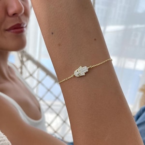 Könnte beinhalten: Ein goldfarbenes Armband mit einem Hamsa-Hand-Anhänger, besetzt mit kleinen, klaren Edelsteinen. Das Armband hat eine feine Kette. Der Anhänger ist der Mittelpunkt und zeigt das detaillierte Design und den Glanz.