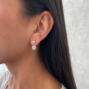 Puede incluir: Primer plano de una oreja adornada con dos pendientes de diamantes. Los pendientes son redondos, con un perno más grande debajo de uno más pequeño. Las joyas brillan. La persona lleva una blusa de encaje blanco. Los pendientes son probablemente de oro blanco o platino.