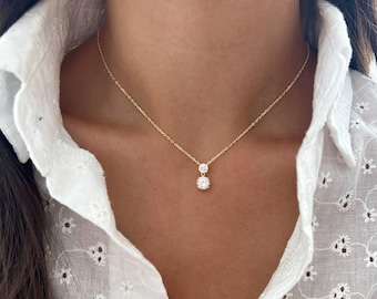 May include: A delicate gold necklace featuring two clear, round gemstones. The necklace is suspended from a fine gold chain and rests against a white, embroidered blouse. The gemstones are set in a classic, minimalist style.