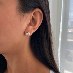 May include: A close-up of an ear with two diamond stud earrings. The earrings have a round and a pear-shaped diamond. The diamonds are set in a gold or silver setting. The person is wearing a white lace top.