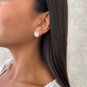 May include: A close-up of a person wearing a pair of pear-shaped diamond stud earrings. The earrings are set in a gold or silver metal. The person is wearing a white lace top.