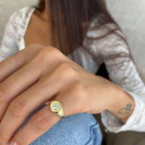 May include: A gold signet ring featuring a circular face with a small, clear gemstone at its center. The ring is worn on a finger, with a light-colored top and blue jeans visible in the background.