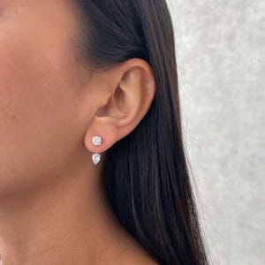 May include: A close-up of a person's ear adorned with two diamond earrings. The top earring is a round, square-cut diamond, and the bottom earring is a pear-shaped diamond. The earrings sparkle against the skin.