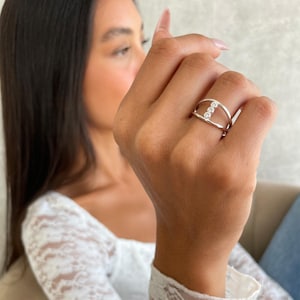 May include: A silver ring with three small, clear stones set in a horizontal line. The ring has a double-band design, with one band above and one below the stones. The ring is displayed on a hand, with a blurred background.