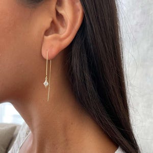 May include: A close-up of a person's ear with a gold threader earring. The earring features a small, diamond-shaped charm with a clear stone in the center. The earring hangs down, with a delicate chain and a straight bar.